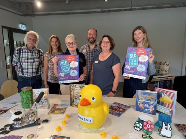 Sechs Personen stehen hinter einem Tisch in einer Bibliothek und präsentieren Plakate für den „SLC SommerLeseClub“. Auf dem Tisch liegen Kinder- und Jugendbücher, eine große gelbe Ente mit der Aufschrift „Mama Berta“, mehrere kleine Badeenten sowie Bastelarbeiten und Werbematerialien. Die Aufnahme vermittelt den Eindruck einer gemeinsamen Auftaktveranstaltung zur Leseförderung und lädt zur Teilnahme am Sommerleseclub ein.