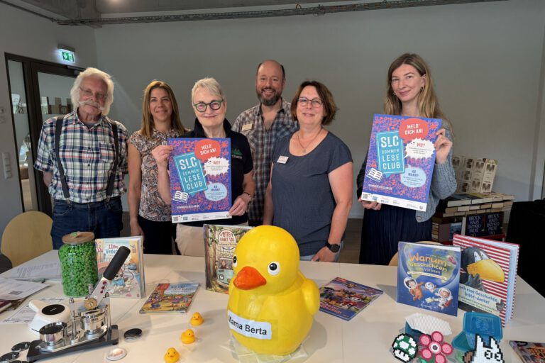 Foto: Stadt Menden Sechs Personen stehen hinter einem Tisch in einer Bibliothek und präsentieren Plakate für den „SLC SommerLeseClub“. Auf dem Tisch liegen Kinder- und Jugendbücher, eine große gelbe Ente mit der Aufschrift „Mama Berta“, mehrere kleine Badeenten sowie Bastelarbeiten und Werbematerialien. Die Aufnahme vermittelt den Eindruck einer gemeinsamen Auftaktveranstaltung zur Leseförderung und lädt zur Teilnahme am Sommerleseclub ein.