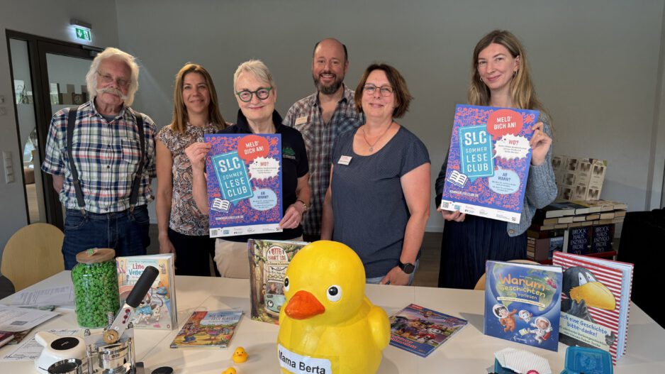 Sechs Personen stehen hinter einem Tisch in einer Bibliothek und präsentieren Plakate für den „SLC SommerLeseClub“. Auf dem Tisch liegen Kinder- und Jugendbücher, eine große gelbe Ente mit der Aufschrift „Mama Berta“, mehrere kleine Badeenten sowie Bastelarbeiten und Werbematerialien. Die Aufnahme vermittelt den Eindruck einer gemeinsamen Auftaktveranstaltung zur Leseförderung und lädt zur Teilnahme am Sommerleseclub ein.