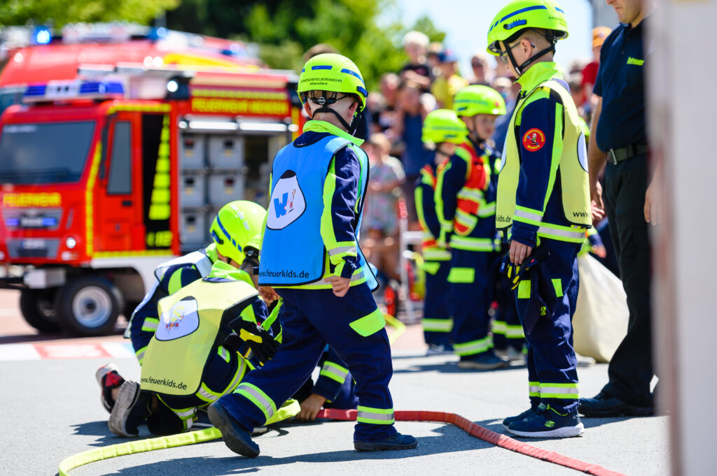 Mehrere Kinder der Kinderfeuerwehr tragen leuchtend gelbe Helme und Schutzwesten und üben mit Feuerwehrschläuchen auf einem abgesperrten Platz. Im Hintergrund steht ein rotes Feuerwehrfahrzeug, während zahlreiche Zuschauer das Geschehen verfolgen. Die Szene vermittelt spielerisch den Feuerwehralltag und die Nachwuchsarbeit der Feuerwehr bei einer öffentlichen Veranstaltung.