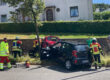 Einsatzkräfte der Feuerwehr und des Rettungsdienstes kümmern sich nach einem Verkehrsunfall um die Beteiligten. Ein schwarzer Kleinwagen und ein rotes Fahrzeug stehen beschädigt am Fahrbahnrand. Mehrere Feuerwehrleute arbeiten an einem der Fahrzeuge, während Einsatzmaterial auf der Straße bereitliegt. Im Hintergrund befinden sich Wohnhäuser und eine Hecke. Die Szene zeigt einen Rettungseinsatz nach einer Kollision im Wohngebiet.