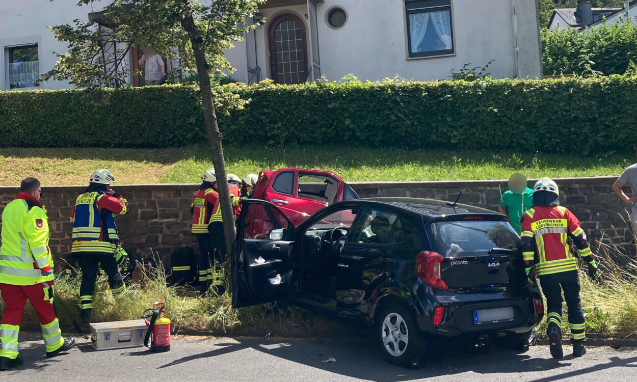 Einsatzkräfte der Feuerwehr und des Rettungsdienstes kümmern sich nach einem Verkehrsunfall um die Beteiligten. Ein schwarzer Kleinwagen und ein rotes Fahrzeug stehen beschädigt am Fahrbahnrand. Mehrere Feuerwehrleute arbeiten an einem der Fahrzeuge, während Einsatzmaterial auf der Straße bereitliegt. Im Hintergrund befinden sich Wohnhäuser und eine Hecke. Die Szene zeigt einen Rettungseinsatz nach einer Kollision im Wohngebiet.