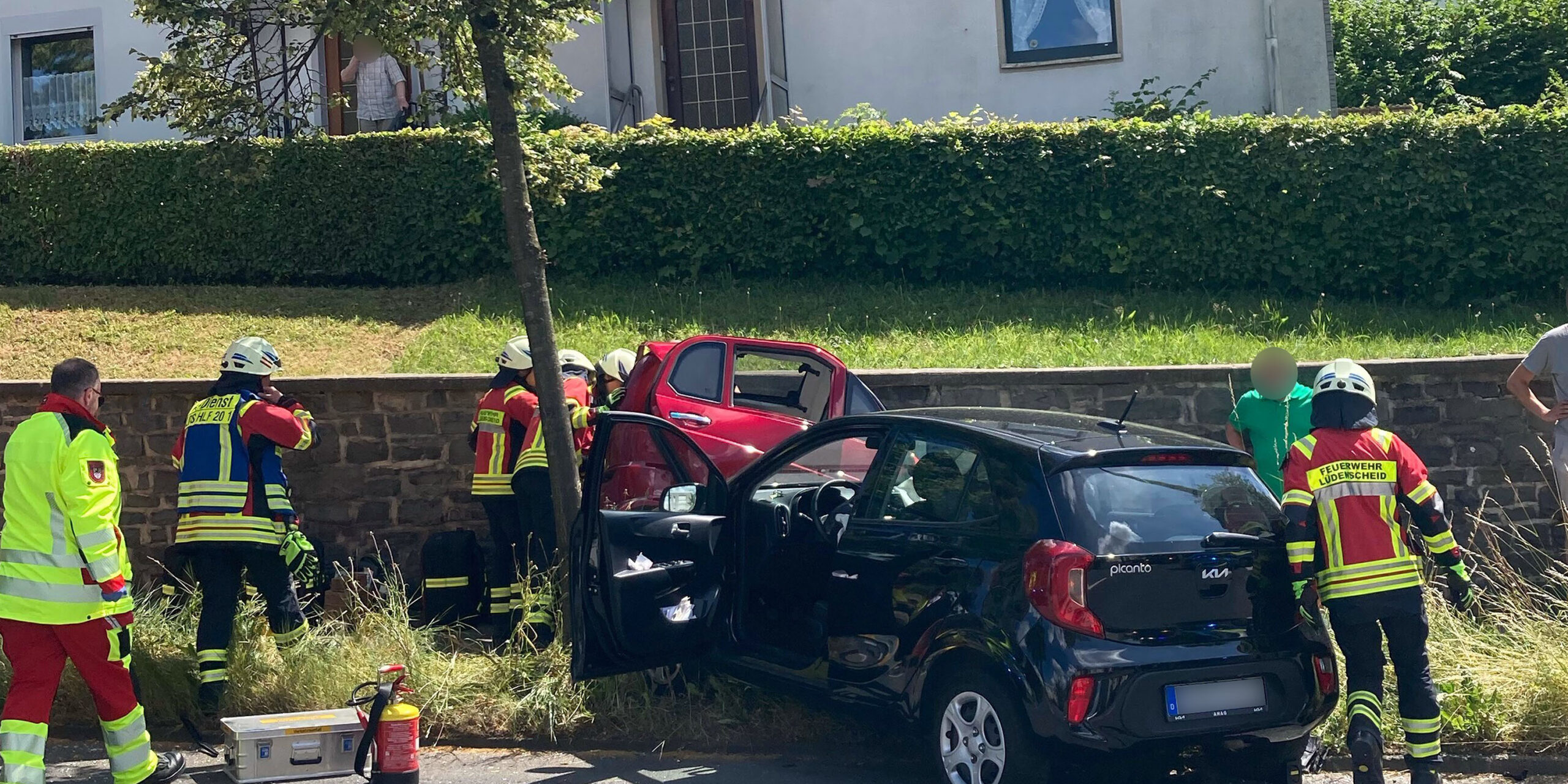 Einsatzkräfte der Feuerwehr und des Rettungsdienstes kümmern sich nach einem Verkehrsunfall um die Beteiligten. Ein schwarzer Kleinwagen und ein rotes Fahrzeug stehen beschädigt am Fahrbahnrand. Mehrere Feuerwehrleute arbeiten an einem der Fahrzeuge, während Einsatzmaterial auf der Straße bereitliegt. Im Hintergrund befinden sich Wohnhäuser und eine Hecke. Die Szene zeigt einen Rettungseinsatz nach einer Kollision im Wohngebiet.
