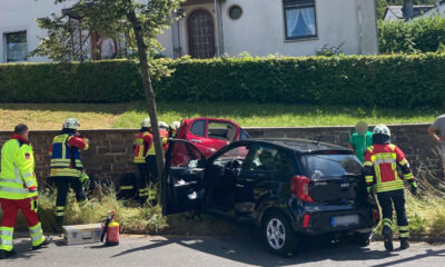 Einsatzkräfte der Feuerwehr und des Rettungsdienstes kümmern sich nach einem Verkehrsunfall um die Beteiligten. Ein schwarzer Kleinwagen und ein rotes Fahrzeug stehen beschädigt am Fahrbahnrand. Mehrere Feuerwehrleute arbeiten an einem der Fahrzeuge, während Einsatzmaterial auf der Straße bereitliegt. Im Hintergrund befinden sich Wohnhäuser und eine Hecke. Die Szene zeigt einen Rettungseinsatz nach einer Kollision im Wohngebiet.