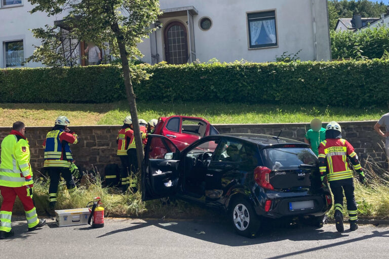 Foto: Polizei MK Einsatzkräfte der Feuerwehr und des Rettungsdienstes kümmern sich nach einem Verkehrsunfall um die Beteiligten. Ein schwarzer Kleinwagen und ein rotes Fahrzeug stehen beschädigt am Fahrbahnrand. Mehrere Feuerwehrleute arbeiten an einem der Fahrzeuge, während Einsatzmaterial auf der Straße bereitliegt. Im Hintergrund befinden sich Wohnhäuser und eine Hecke. Die Szene zeigt einen Rettungseinsatz nach einer Kollision im Wohngebiet.