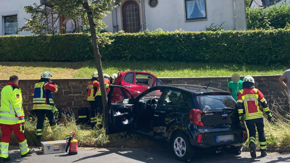 Einsatzkräfte der Feuerwehr und des Rettungsdienstes kümmern sich nach einem Verkehrsunfall um die Beteiligten. Ein schwarzer Kleinwagen und ein rotes Fahrzeug stehen beschädigt am Fahrbahnrand. Mehrere Feuerwehrleute arbeiten an einem der Fahrzeuge, während Einsatzmaterial auf der Straße bereitliegt. Im Hintergrund befinden sich Wohnhäuser und eine Hecke. Die Szene zeigt einen Rettungseinsatz nach einer Kollision im Wohngebiet.