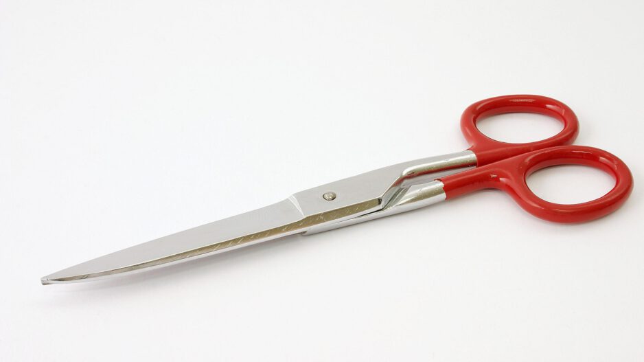 Eine silberne Schere mit roten Kunststoffgriffen liegt geschlossen auf einem weißen Hintergrund. Die Aufnahme zeigt das Schneidwerkzeug schräg von oben, wobei die langen Metallklingen nach links und die Griffe nach rechts weisen. Durch den neutralen Hintergrund steht die Schere klar im Mittelpunkt des Bildes.