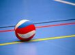 Ein rot-weiß-blauer Volleyball liegt auf dem blauen Hallenboden einer Sporthalle. Farbige Spielfeldmarkierungen in Gelb, Rot und Schwarz verlaufen um den Ball und betonen die geometrische Anordnung der Linien. Das Bild symbolisiert Hallenvolleyball und den Beginn oder das Ende eines Spiels oder Trainings.