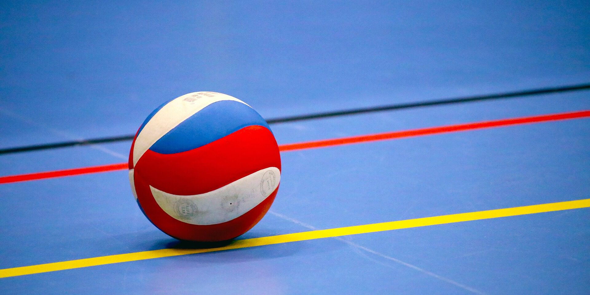Ein rot-weiß-blauer Volleyball liegt auf dem blauen Hallenboden einer Sporthalle. Farbige Spielfeldmarkierungen in Gelb, Rot und Schwarz verlaufen um den Ball und betonen die geometrische Anordnung der Linien. Das Bild symbolisiert Hallenvolleyball und den Beginn oder das Ende eines Spiels oder Trainings.