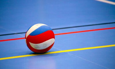 Ein rot-weiß-blauer Volleyball liegt auf dem blauen Hallenboden einer Sporthalle. Farbige Spielfeldmarkierungen in Gelb, Rot und Schwarz verlaufen um den Ball und betonen die geometrische Anordnung der Linien. Das Bild symbolisiert Hallenvolleyball und den Beginn oder das Ende eines Spiels oder Trainings.