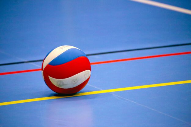 Foto: Tania Van den Berghen/Pixabay Ein rot-weiß-blauer Volleyball liegt auf dem blauen Hallenboden einer Sporthalle. Farbige Spielfeldmarkierungen in Gelb, Rot und Schwarz verlaufen um den Ball und betonen die geometrische Anordnung der Linien. Das Bild symbolisiert Hallenvolleyball und den Beginn oder das Ende eines Spiels oder Trainings.