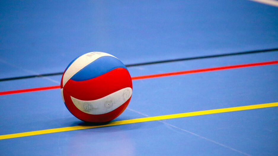 Ein rot-weiß-blauer Volleyball liegt auf dem blauen Hallenboden einer Sporthalle. Farbige Spielfeldmarkierungen in Gelb, Rot und Schwarz verlaufen um den Ball und betonen die geometrische Anordnung der Linien. Das Bild symbolisiert Hallenvolleyball und den Beginn oder das Ende eines Spiels oder Trainings.