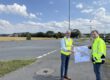 Zwei Männer in leuchtenden Warnwesten stehen an einer Straßenkreuzung und halten gemeinsam einen Plan für die Umgestaltung des Verkehrsbereichs. Im Hintergrund sind Felder, Wohnhäuser und Wegweiser zu sehen.