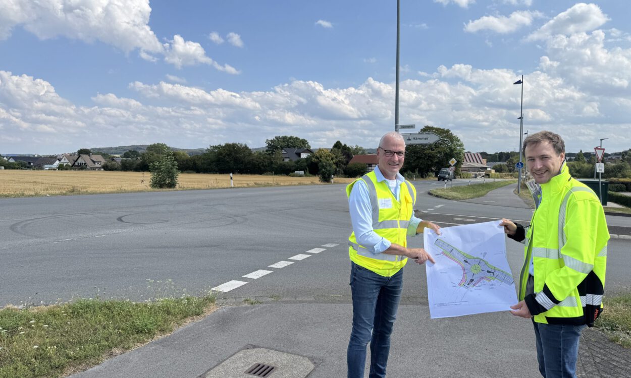 Zwei Männer in leuchtenden Warnwesten stehen an einer Straßenkreuzung und halten gemeinsam einen Plan für die Umgestaltung des Verkehrsbereichs. Im Hintergrund sind Felder, Wohnhäuser und Wegweiser zu sehen.