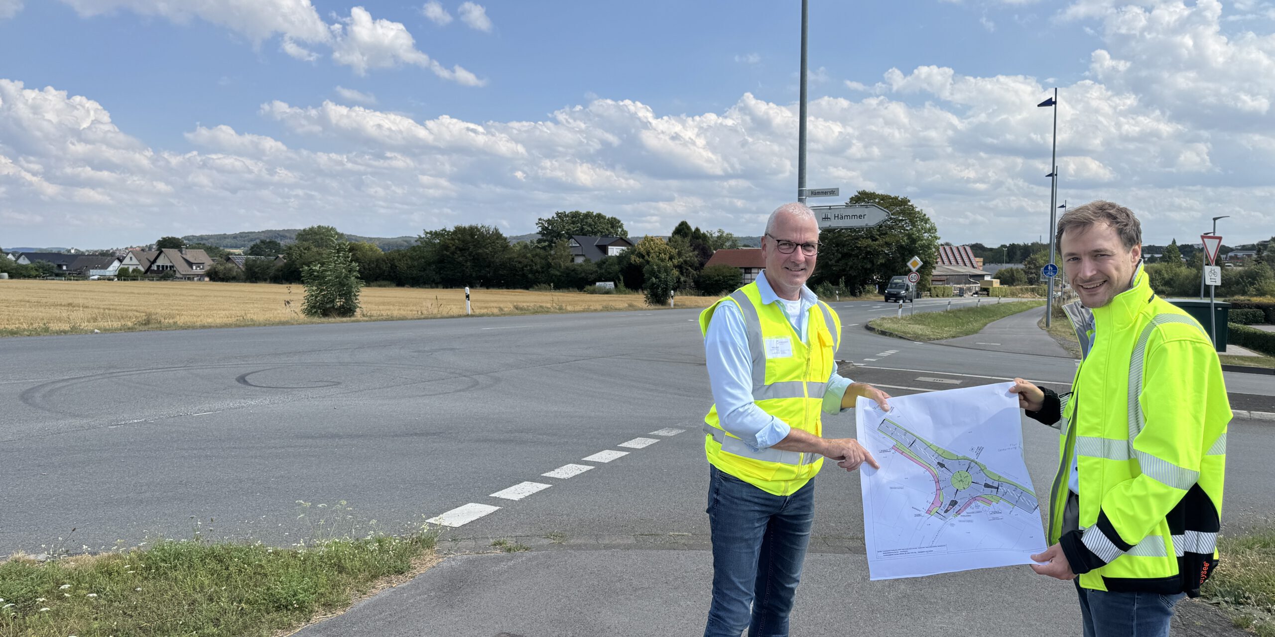 Zwei Männer in leuchtenden Warnwesten stehen an einer Straßenkreuzung und halten gemeinsam einen Plan für die Umgestaltung des Verkehrsbereichs. Im Hintergrund sind Felder, Wohnhäuser und Wegweiser zu sehen.