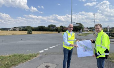 Zwei Männer in leuchtenden Warnwesten stehen an einer Straßenkreuzung und halten gemeinsam einen Plan für die Umgestaltung des Verkehrsbereichs. Im Hintergrund sind Felder, Wohnhäuser und Wegweiser zu sehen.