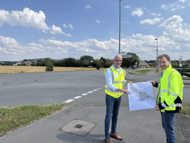 Zwei Männer in leuchtenden Warnwesten stehen an einer Straßenkreuzung und halten gemeinsam einen Plan für die Umgestaltung des Verkehrsbereichs. Im Hintergrund sind Felder, Wohnhäuser und Wegweiser zu sehen.