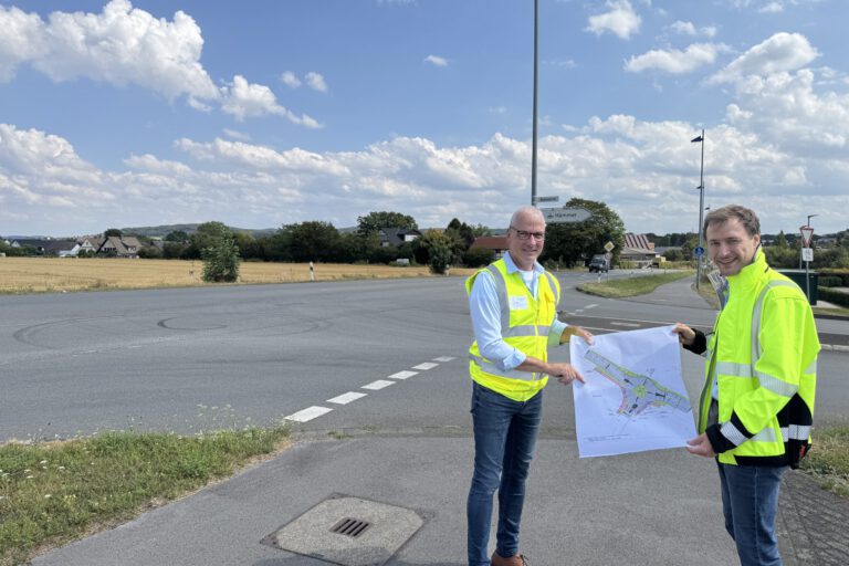 Zwei Männer in leuchtenden Warnwesten stehen an einer Straßenkreuzung und halten gemeinsam einen Plan für die Umgestaltung des Verkehrsbereichs. Im Hintergrund sind Felder, Wohnhäuser und Wegweiser zu sehen.