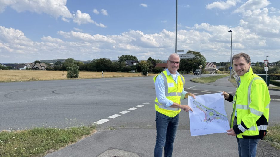 Zwei Männer in leuchtenden Warnwesten stehen an einer Straßenkreuzung und halten gemeinsam einen Plan für die Umgestaltung des Verkehrsbereichs. Im Hintergrund sind Felder, Wohnhäuser und Wegweiser zu sehen.