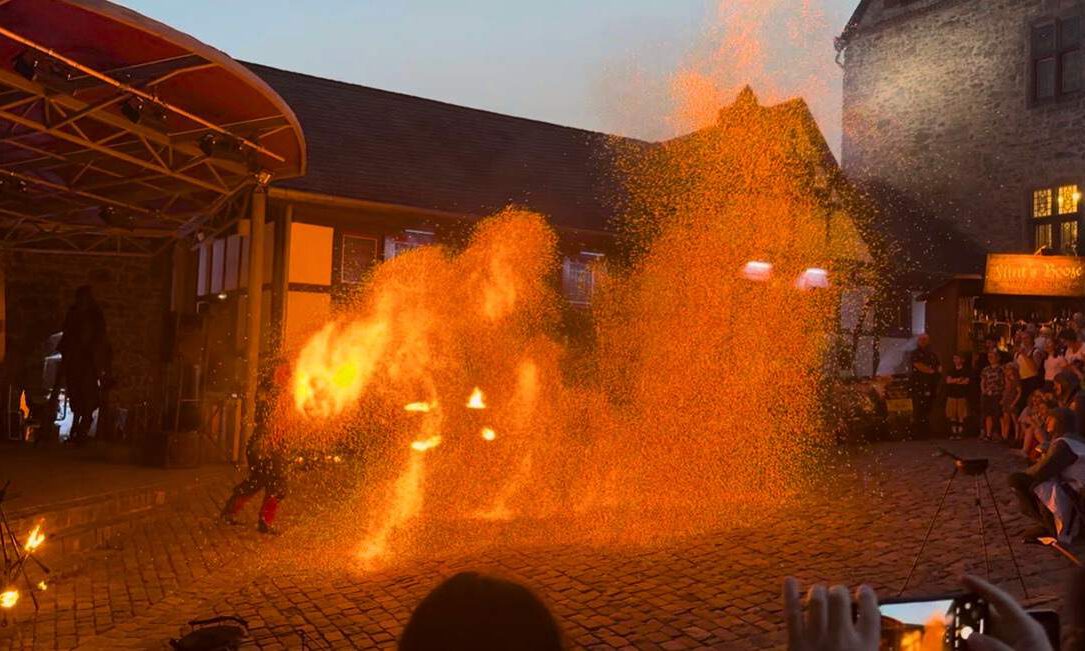 Eine Feuershow findet am Abend auf einem historischen Burghof statt. Vor einer Bühne erzeugt ein Künstler mit brennenden Requisiten und Funken spektakuläre Flammenbilder, die den Platz in warmes orangefarbenes Licht tauchen. Zahlreiche Zuschauer verfolgen die Vorführung rund um den Platz, einige halten den Moment mit ihren Smartphones fest.