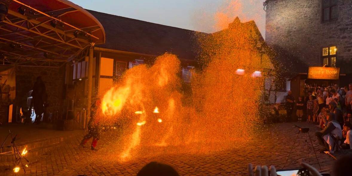 Eine Feuershow findet am Abend auf einem historischen Burghof statt. Vor einer Bühne erzeugt ein Künstler mit brennenden Requisiten und Funken spektakuläre Flammenbilder, die den Platz in warmes orangefarbenes Licht tauchen. Zahlreiche Zuschauer verfolgen die Vorführung rund um den Platz, einige halten den Moment mit ihren Smartphones fest.
