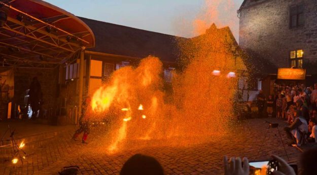 Eine Feuershow findet am Abend auf einem historischen Burghof statt. Vor einer Bühne erzeugt ein Künstler mit brennenden Requisiten und Funken spektakuläre Flammenbilder, die den Platz in warmes orangefarbenes Licht tauchen. Zahlreiche Zuschauer verfolgen die Vorführung rund um den Platz, einige halten den Moment mit ihren Smartphones fest.