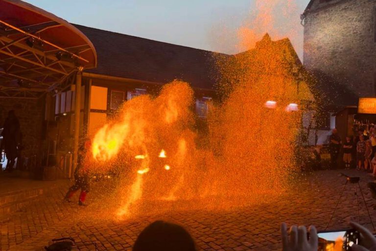 Foto: Eileen Bräuniger/Märkischer Kreis Eine Feuershow findet am Abend auf einem historischen Burghof statt. Vor einer Bühne erzeugt ein Künstler mit brennenden Requisiten und Funken spektakuläre Flammenbilder, die den Platz in warmes orangefarbenes Licht tauchen. Zahlreiche Zuschauer verfolgen die Vorführung rund um den Platz, einige halten den Moment mit ihren Smartphones fest.