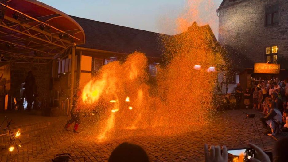 Eine Feuershow findet am Abend auf einem historischen Burghof statt. Vor einer Bühne erzeugt ein Künstler mit brennenden Requisiten und Funken spektakuläre Flammenbilder, die den Platz in warmes orangefarbenes Licht tauchen. Zahlreiche Zuschauer verfolgen die Vorführung rund um den Platz, einige halten den Moment mit ihren Smartphones fest.