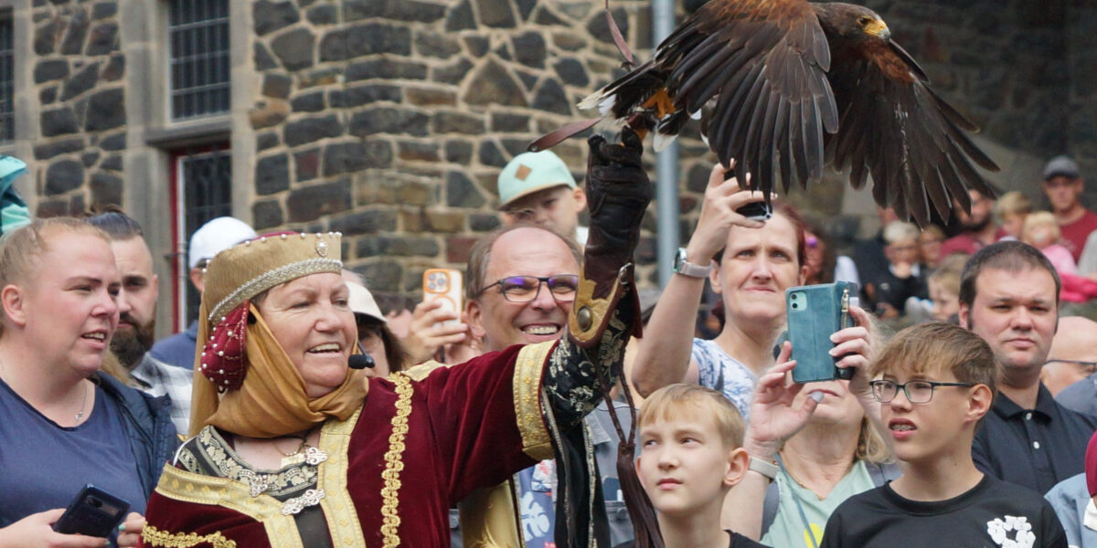 Eine Frau in mittelalterlich anmutender Gewandung präsentiert einen Greifvogel, der mit ausgebreiteten Flügeln auf ihrem behandschuhten Arm sitzt. Rundherum verfolgt eine große Menschenmenge die Vorführung, viele halten Smartphones hoch oder blicken gespannt auf den Vogel. Im Hintergrund ist die Natursteinfassade einer Burg oder historischen Anlage zu sehen.