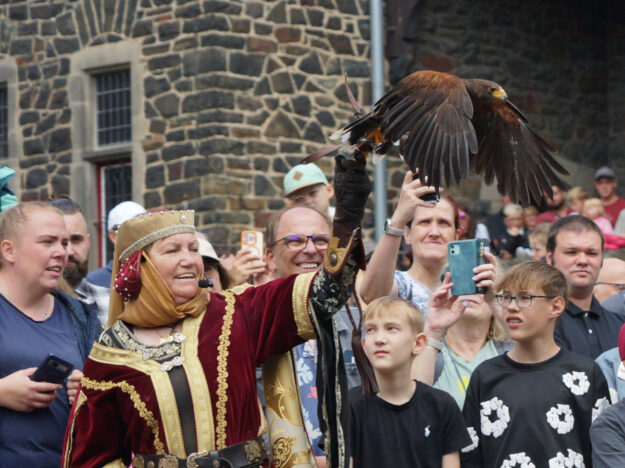 Foto: Britta Gerstendorf/Märkischer Kreis Eine Frau in mittelalterlich anmutender Gewandung präsentiert einen Greifvogel, der mit ausgebreiteten Flügeln auf ihrem behandschuhten Arm sitzt. Rundherum verfolgt eine große Menschenmenge die Vorführung, viele halten Smartphones hoch oder blicken gespannt auf den Vogel. Im Hintergrund ist die Natursteinfassade einer Burg oder historischen Anlage zu sehen.