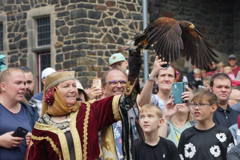 Eine Frau in mittelalterlich anmutender Gewandung präsentiert einen Greifvogel, der mit ausgebreiteten Flügeln auf ihrem behandschuhten Arm sitzt. Rundherum verfolgt eine große Menschenmenge die Vorführung, viele halten Smartphones hoch oder blicken gespannt auf den Vogel. Im Hintergrund ist die Natursteinfassade einer Burg oder historischen Anlage zu sehen.