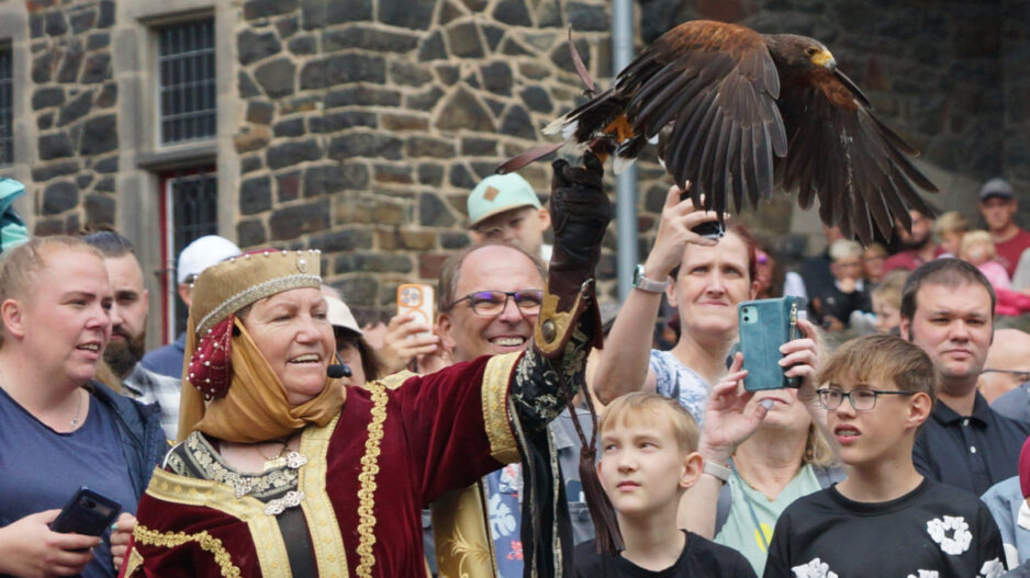 Eine Frau in mittelalterlich anmutender Gewandung präsentiert einen Greifvogel, der mit ausgebreiteten Flügeln auf ihrem behandschuhten Arm sitzt. Rundherum verfolgt eine große Menschenmenge die Vorführung, viele halten Smartphones hoch oder blicken gespannt auf den Vogel. Im Hintergrund ist die Natursteinfassade einer Burg oder historischen Anlage zu sehen.