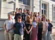 Eine Gruppe von Auszubildenden und ihren Begleitpersonen steht auf den Treppenstufen vor dem Eingang eines historischen Verwaltungsgebäudes für ein gemeinsames Gruppenfoto. Die jungen Erwachsenen sind in mehreren Reihen angeordnet und blicken in die Kamera. Die Aufnahme entstand bei sonnigem Wetter und vermittelt den Eindruck eines offiziellen Auftakts oder Begrüßungstermins zum Ausbildungsbeginn.