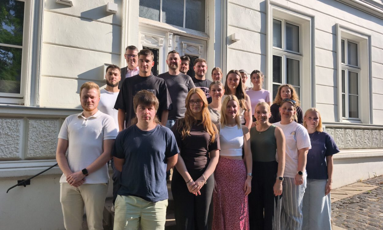 Eine Gruppe von Auszubildenden und ihren Begleitpersonen steht auf den Treppenstufen vor dem Eingang eines historischen Verwaltungsgebäudes für ein gemeinsames Gruppenfoto. Die jungen Erwachsenen sind in mehreren Reihen angeordnet und blicken in die Kamera. Die Aufnahme entstand bei sonnigem Wetter und vermittelt den Eindruck eines offiziellen Auftakts oder Begrüßungstermins zum Ausbildungsbeginn.