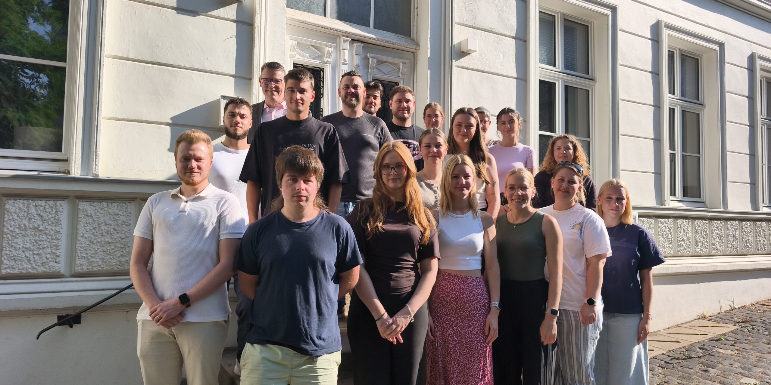 Eine Gruppe von Auszubildenden und ihren Begleitpersonen steht auf den Treppenstufen vor dem Eingang eines historischen Verwaltungsgebäudes für ein gemeinsames Gruppenfoto. Die jungen Erwachsenen sind in mehreren Reihen angeordnet und blicken in die Kamera. Die Aufnahme entstand bei sonnigem Wetter und vermittelt den Eindruck eines offiziellen Auftakts oder Begrüßungstermins zum Ausbildungsbeginn.