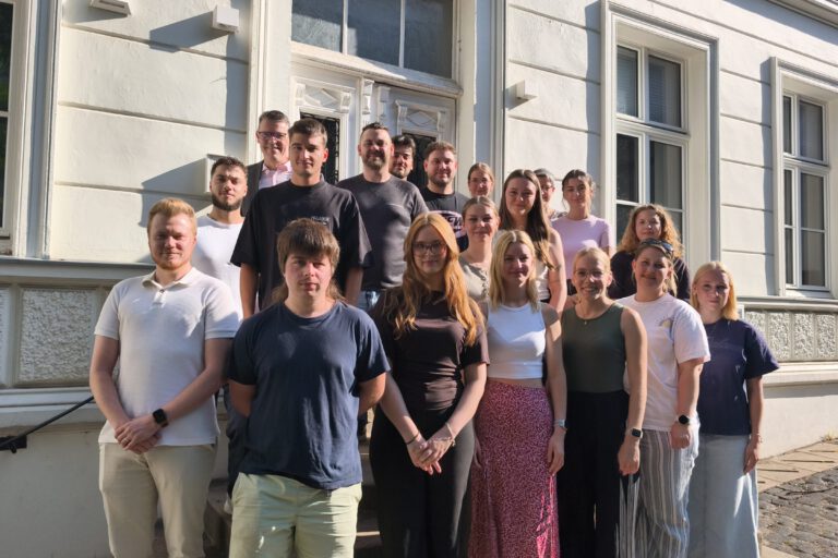 Eine Gruppe von Auszubildenden und ihren Begleitpersonen steht auf den Treppenstufen vor dem Eingang eines historischen Verwaltungsgebäudes für ein gemeinsames Gruppenfoto. Die jungen Erwachsenen sind in mehreren Reihen angeordnet und blicken in die Kamera. Die Aufnahme entstand bei sonnigem Wetter und vermittelt den Eindruck eines offiziellen Auftakts oder Begrüßungstermins zum Ausbildungsbeginn.