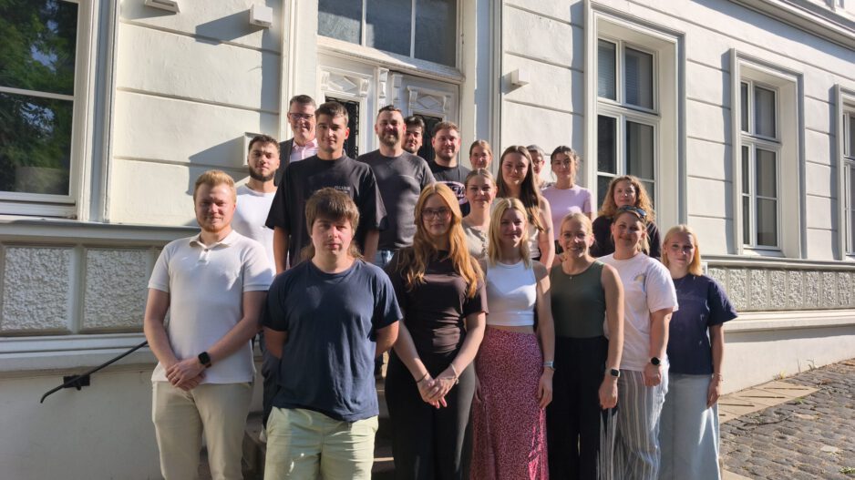 Eine Gruppe von Auszubildenden und ihren Begleitpersonen steht auf den Treppenstufen vor dem Eingang eines historischen Verwaltungsgebäudes für ein gemeinsames Gruppenfoto. Die jungen Erwachsenen sind in mehreren Reihen angeordnet und blicken in die Kamera. Die Aufnahme entstand bei sonnigem Wetter und vermittelt den Eindruck eines offiziellen Auftakts oder Begrüßungstermins zum Ausbildungsbeginn.
