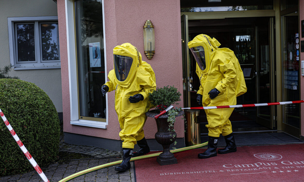 Zwei Einsatzkräfte in gelben Chemikalienschutzanzügen verlassen den Eingang eines Hotels und bewegen sich über den abgesperrten Eingangsbereich. Feuerwehrschläuche und Absperrband deuten auf einen Gefahrguteinsatz hin. Die Aufnahme zeigt den Einsatz unter besonderen Schutzmaßnahmen vor dem Gebäude.