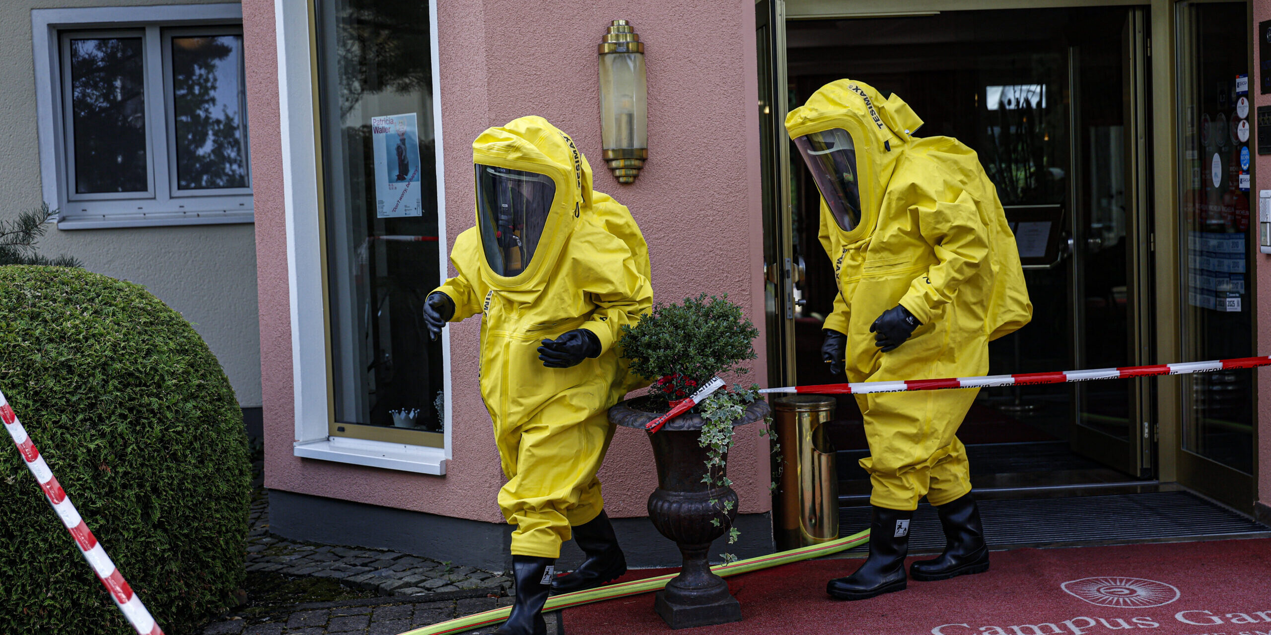 Zwei Einsatzkräfte in gelben Chemikalienschutzanzügen verlassen den Eingang eines Hotels und bewegen sich über den abgesperrten Eingangsbereich. Feuerwehrschläuche und Absperrband deuten auf einen Gefahrguteinsatz hin. Die Aufnahme zeigt den Einsatz unter besonderen Schutzmaßnahmen vor dem Gebäude.