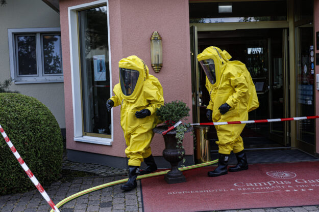 Foto: Dennis Echtermann Zwei Einsatzkräfte in gelben Chemikalienschutzanzügen verlassen den Eingang eines Hotels und bewegen sich über den abgesperrten Eingangsbereich. Feuerwehrschläuche und Absperrband deuten auf einen Gefahrguteinsatz hin. Die Aufnahme zeigt den Einsatz unter besonderen Schutzmaßnahmen vor dem Gebäude.