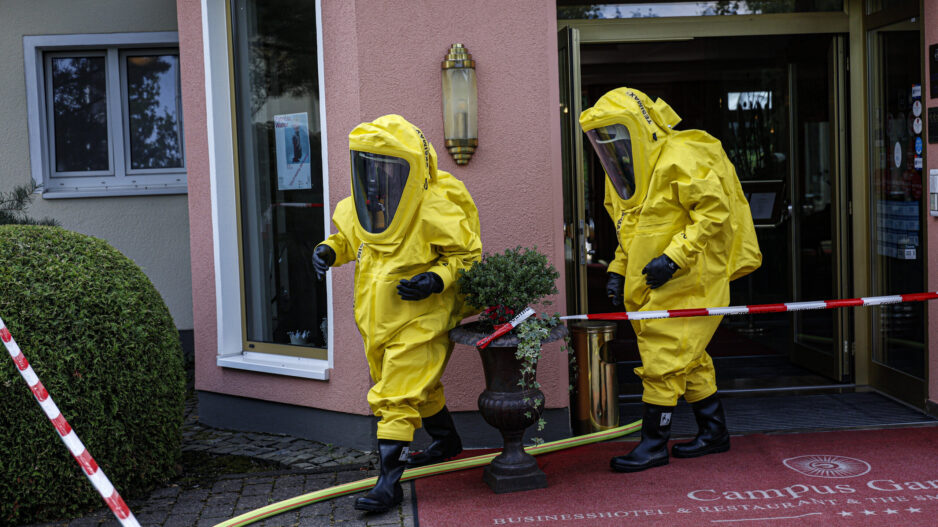Zwei Einsatzkräfte in gelben Chemikalienschutzanzügen verlassen den Eingang eines Hotels und bewegen sich über den abgesperrten Eingangsbereich. Feuerwehrschläuche und Absperrband deuten auf einen Gefahrguteinsatz hin. Die Aufnahme zeigt den Einsatz unter besonderen Schutzmaßnahmen vor dem Gebäude.