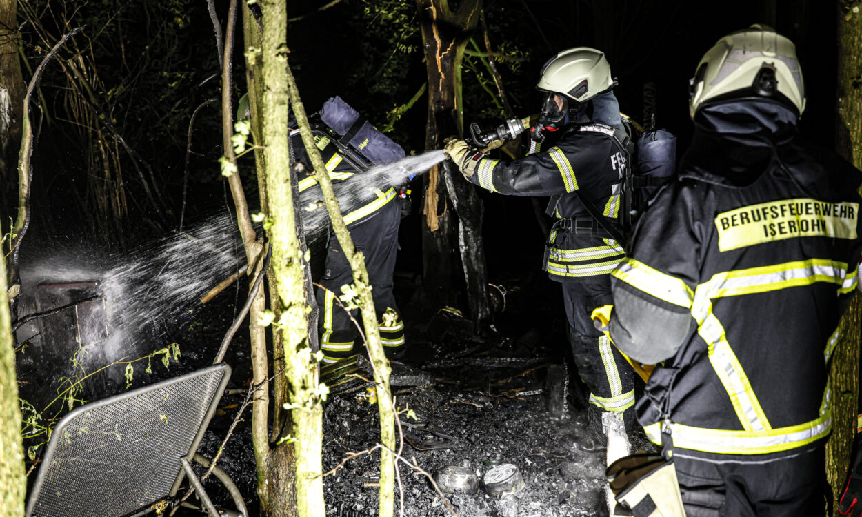 Mehrere Einsatzkräfte der Berufsfeuerwehr löschen nachts in einem Waldstück die Überreste eines Brandes. Ein Feuerwehrmann richtet einen Wasserstrahl auf verkohltes Material, während weitere Einsatzkräfte in Schutzkleidung und mit Atemschutzgeräten unterstützen.
