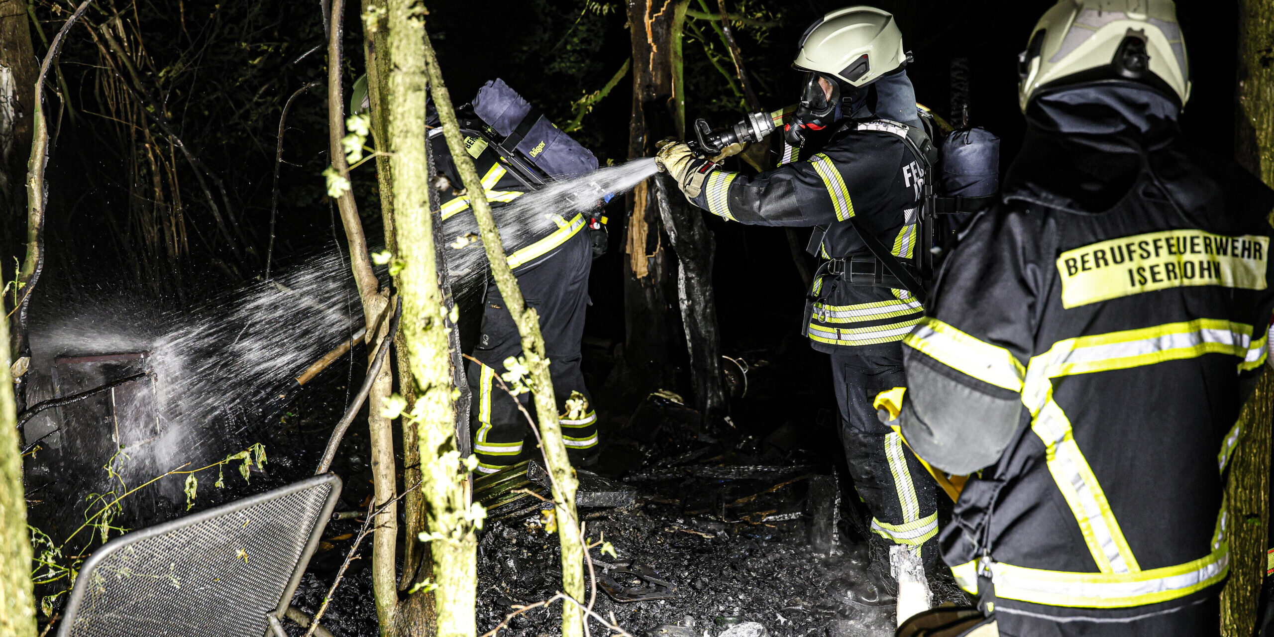 Mehrere Einsatzkräfte der Berufsfeuerwehr löschen nachts in einem Waldstück die Überreste eines Brandes. Ein Feuerwehrmann richtet einen Wasserstrahl auf verkohltes Material, während weitere Einsatzkräfte in Schutzkleidung und mit Atemschutzgeräten unterstützen.