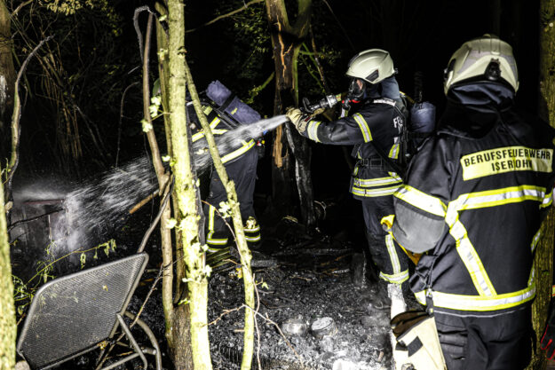 Mehrere Einsatzkräfte der Berufsfeuerwehr löschen nachts in einem Waldstück die Überreste eines Brandes. Ein Feuerwehrmann richtet einen Wasserstrahl auf verkohltes Material, während weitere Einsatzkräfte in Schutzkleidung und mit Atemschutzgeräten unterstützen.