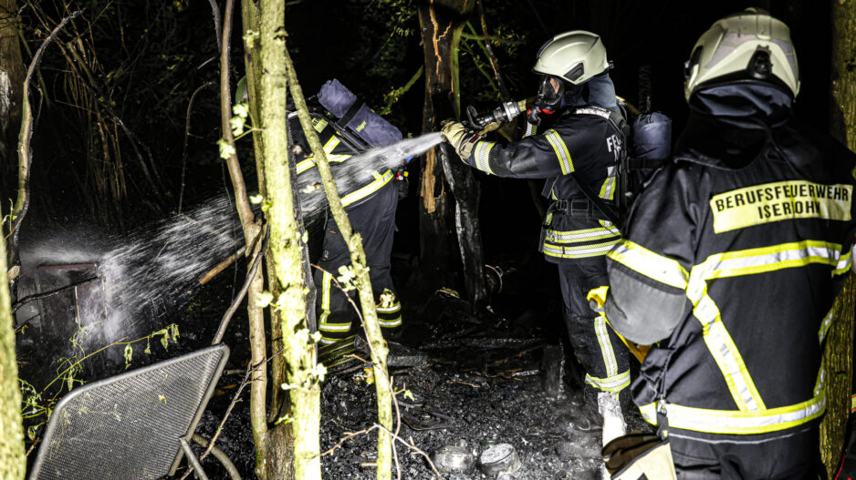 Mehrere Einsatzkräfte der Berufsfeuerwehr löschen nachts in einem Waldstück die Überreste eines Brandes. Ein Feuerwehrmann richtet einen Wasserstrahl auf verkohltes Material, während weitere Einsatzkräfte in Schutzkleidung und mit Atemschutzgeräten unterstützen.