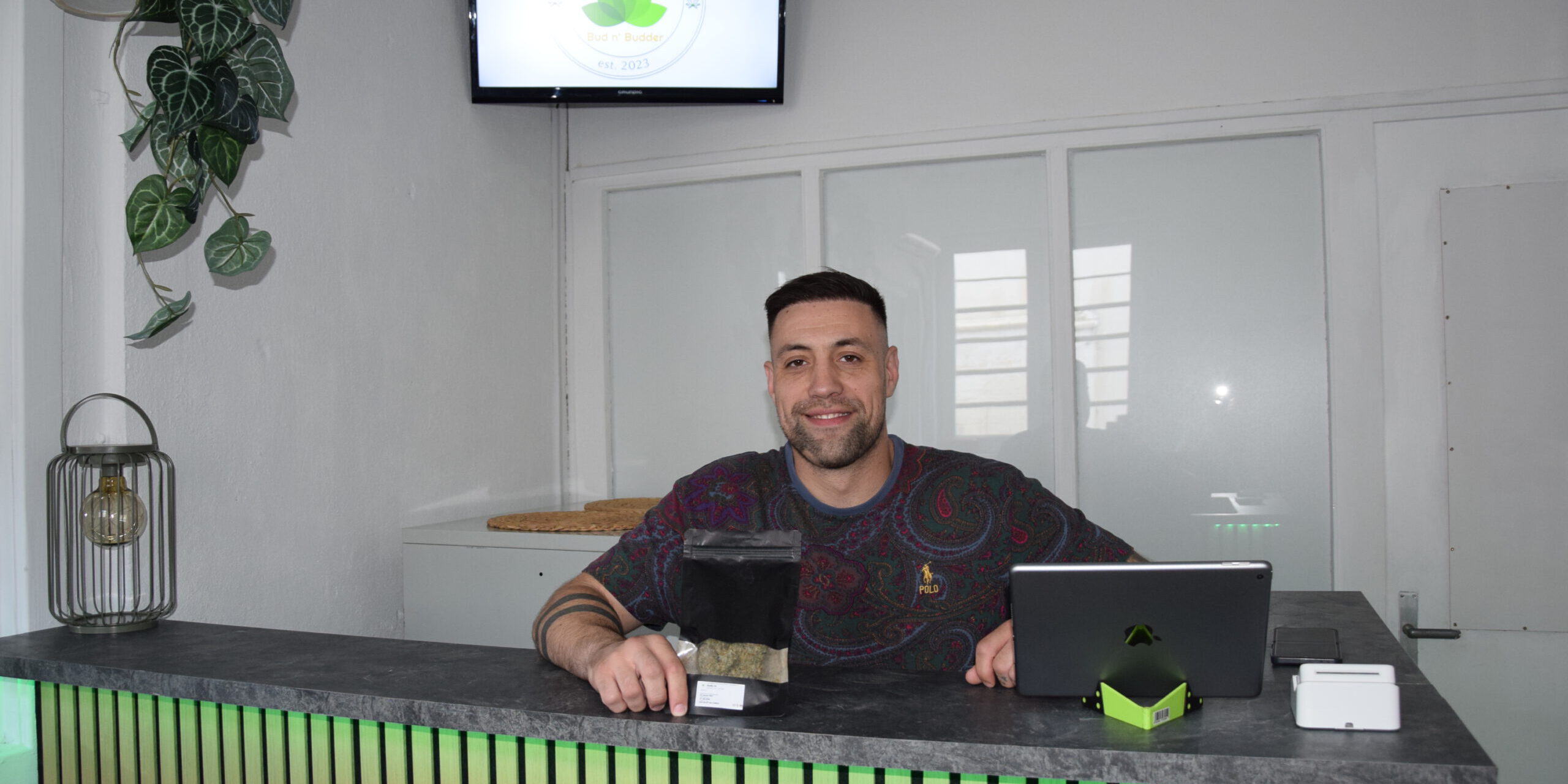 Ein Mann sitzt lächelnd hinter einer Empfangstheke in einem hellen Geschäftsraum. Vor ihm stehen ein Tablet und eine schwarze Produktverpackung. Auf einem Bildschirm an der Wand ist das Logo eines „Cannabis Social Club“ zu sehen. Die Theke ist mit Holzlamellen gestaltet und grün beleuchtet.
