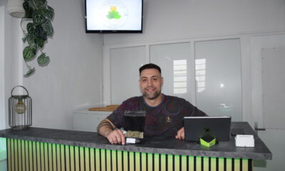 Ein Mann sitzt lächelnd hinter einer Empfangstheke in einem hellen Geschäftsraum. Vor ihm stehen ein Tablet und eine schwarze Produktverpackung. Auf einem Bildschirm an der Wand ist das Logo eines „Cannabis Social Club“ zu sehen. Die Theke ist mit Holzlamellen gestaltet und grün beleuchtet.