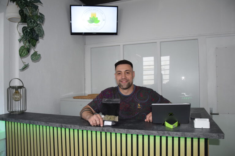 Ein Mann sitzt lächelnd hinter einer Empfangstheke in einem hellen Geschäftsraum. Vor ihm stehen ein Tablet und eine schwarze Produktverpackung. Auf einem Bildschirm an der Wand ist das Logo eines „Cannabis Social Club“ zu sehen. Die Theke ist mit Holzlamellen gestaltet und grün beleuchtet.