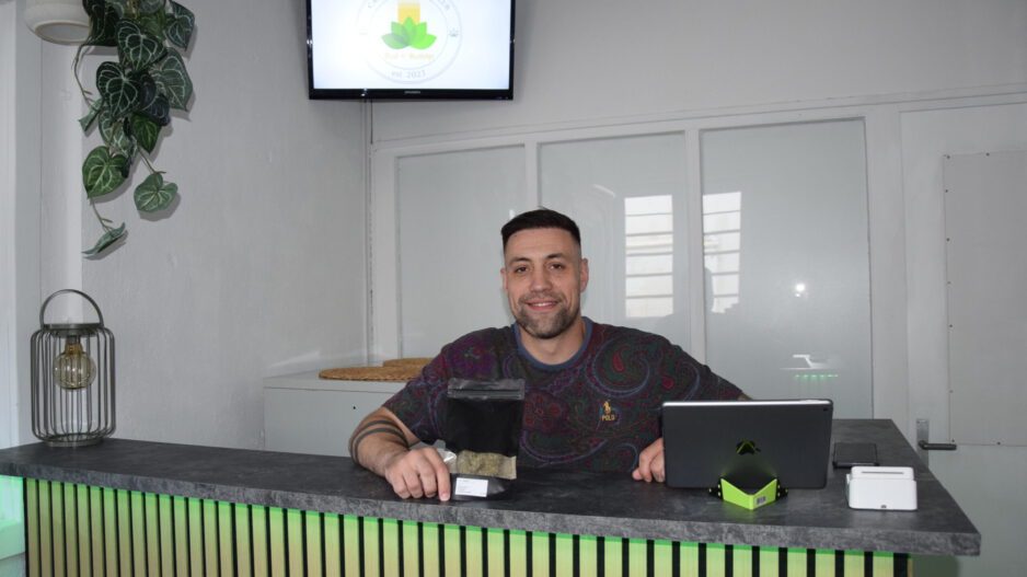 Ein Mann sitzt lächelnd hinter einer Empfangstheke in einem hellen Geschäftsraum. Vor ihm stehen ein Tablet und eine schwarze Produktverpackung. Auf einem Bildschirm an der Wand ist das Logo eines „Cannabis Social Club“ zu sehen. Die Theke ist mit Holzlamellen gestaltet und grün beleuchtet.