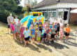 Eine große Gruppe von Kindern und mehreren Erwachsenen steht bei sonnigem Wetter vor einer bunt gelb-blauen Hüpfburg. Die Kinder lächeln in die Kamera und tragen sommerliche Kleidung. Im Hintergrund befindet sich ein Gebäude mit großen Fenstern, umgeben von Bäumen und Sträuchern.